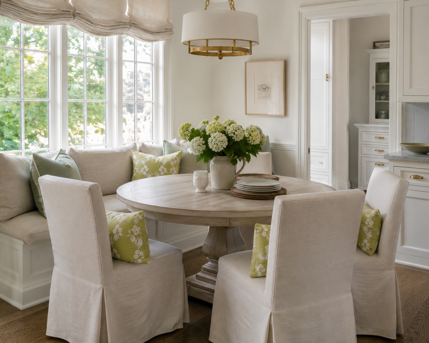 A bright, elegant dining room featuring a round light wood dining table surrounded by four cream slipcovered chairs with green floral accent pillows. A matching cream banquette is positioned along the left wall under a large multi-pane window, decorated with green and cream pillows and a central vase of white hydrangeas.